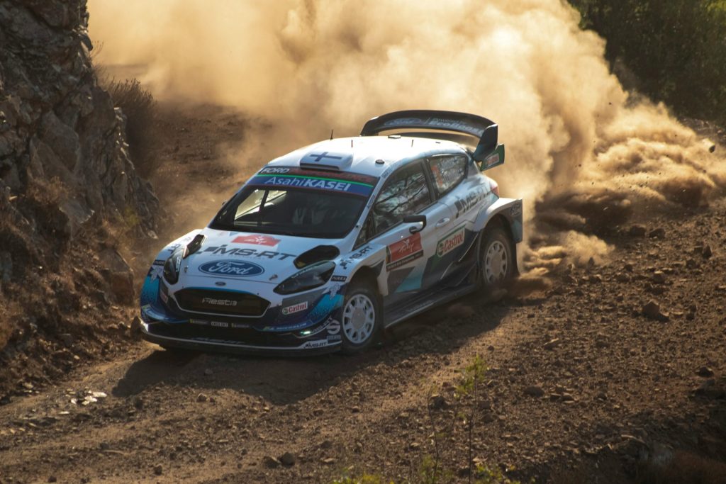 El auto de rally avanza por una curva de un camino de tierra polvorienta, demostrando velocidad y control en una carrera todoterreno al aire libre.